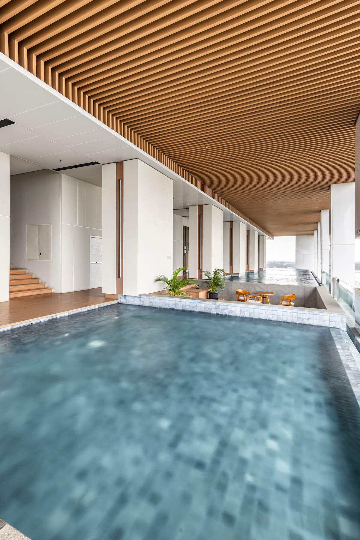 An infinity pool extends along a sleek, modern space, bordered by large windows allowing natural light. Wooden ceiling beams enhance the contemporary design, while comfortable seating is arranged nearby, offering a tranquil spot to relax. The city view is visible in the background.