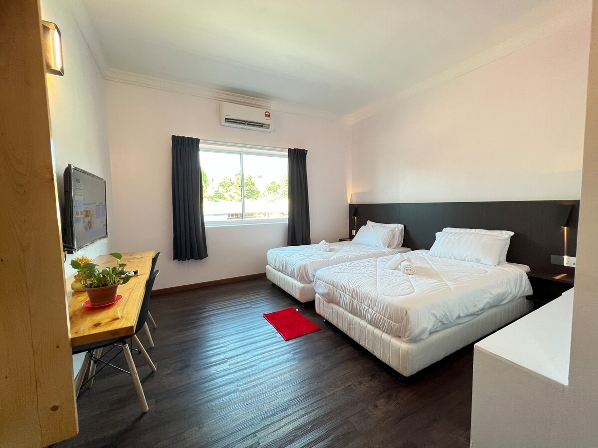 A spacious guest suite features two neatly arranged beds and a wooden dining table with chairs. A flat-screen television is mounted on the wall opposite the beds. Natural light is provided by a large window, accompanied by dark curtains for privacy.