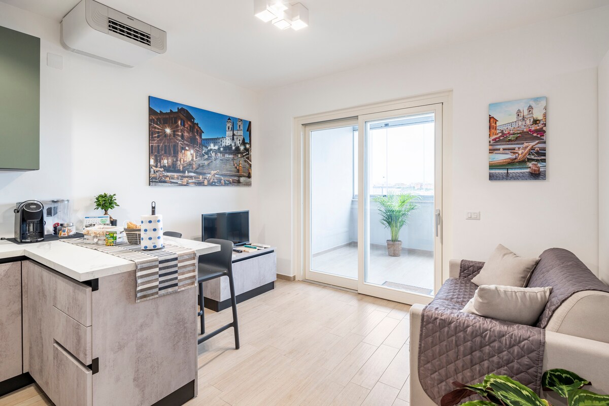 The living area showcases a comfortable sofa next to a small television and a kitchenette featuring modern appliances. A sliding glass door leads to the closed balcony, allowing natural light to fill the space. Artwork depicting city scenes adorns the walls.