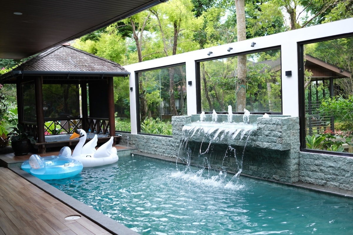 A private pool features a calming water fountain with a gentle flow, surrounded by lush greenery. Inflatable swan floats are placed in the water. A gazebo is visible nearby, offering shaded seating. Large glass windows provide a view of the vibrant garden, enhancing the tranquil setting.