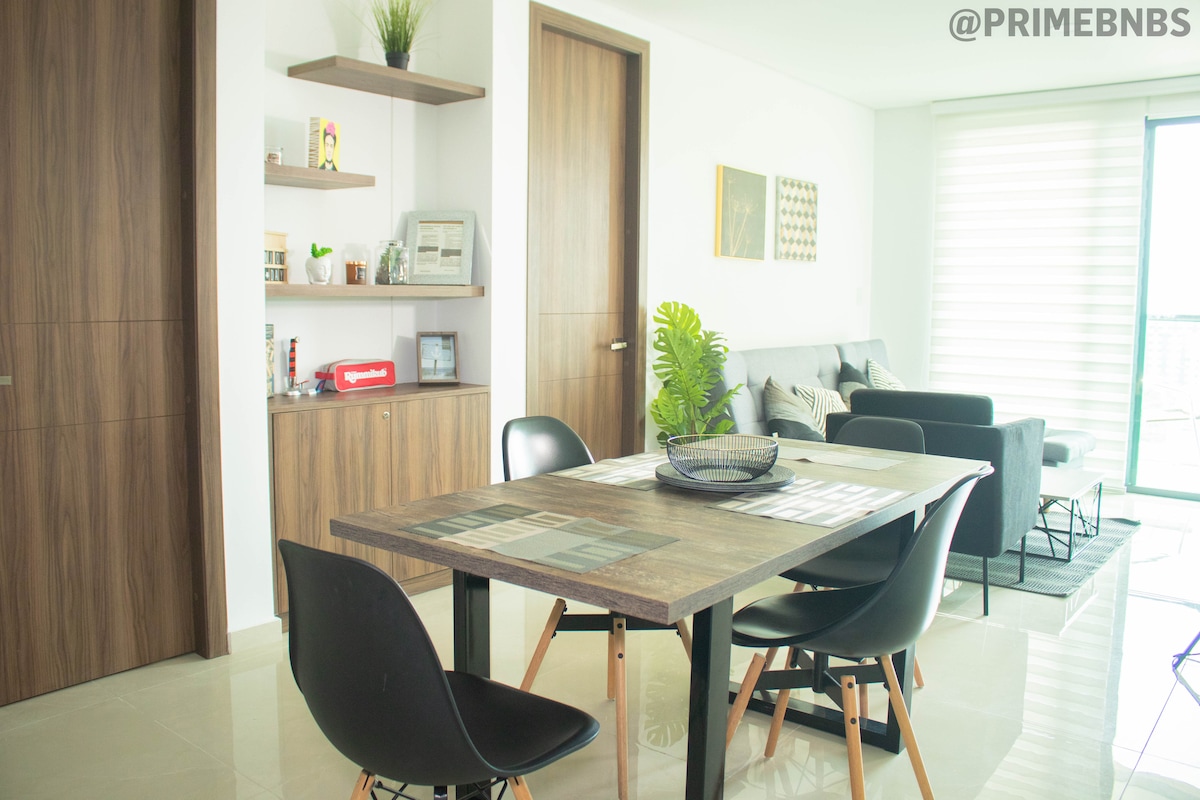 A bright dining area is presented, featuring a wooden table surrounded by black chairs. A shelf with decorative items and a small kitchen appliance is visible in the background. Natural light enters through large glass doors, enhancing the open and airy feel of the space.