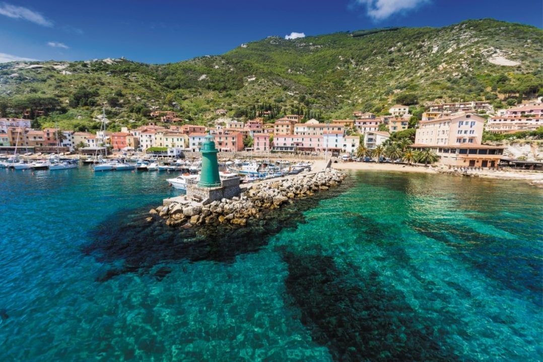 A scenic view of Giglio Porto features a vibrant waterfront lined with colorful buildings. A lighthouse stands on a stone pier, surrounded by clear blue waters that reveal the rocky seabed. The backdrop is dominated by lush green hills under a clear sky.