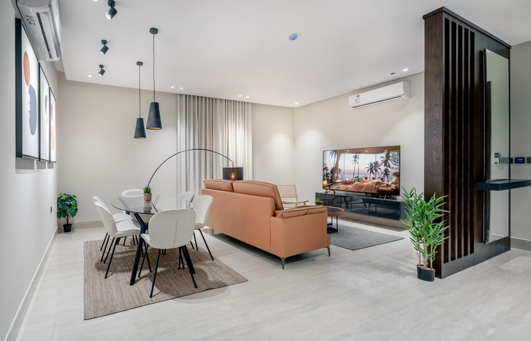 A modern living area features a spacious brown sofa positioned across from a large television. A dining table with white chairs is set against a neutral backdrop. Soft lighting is provided by black pendant fixtures, and decorative plants add a touch of greenery.