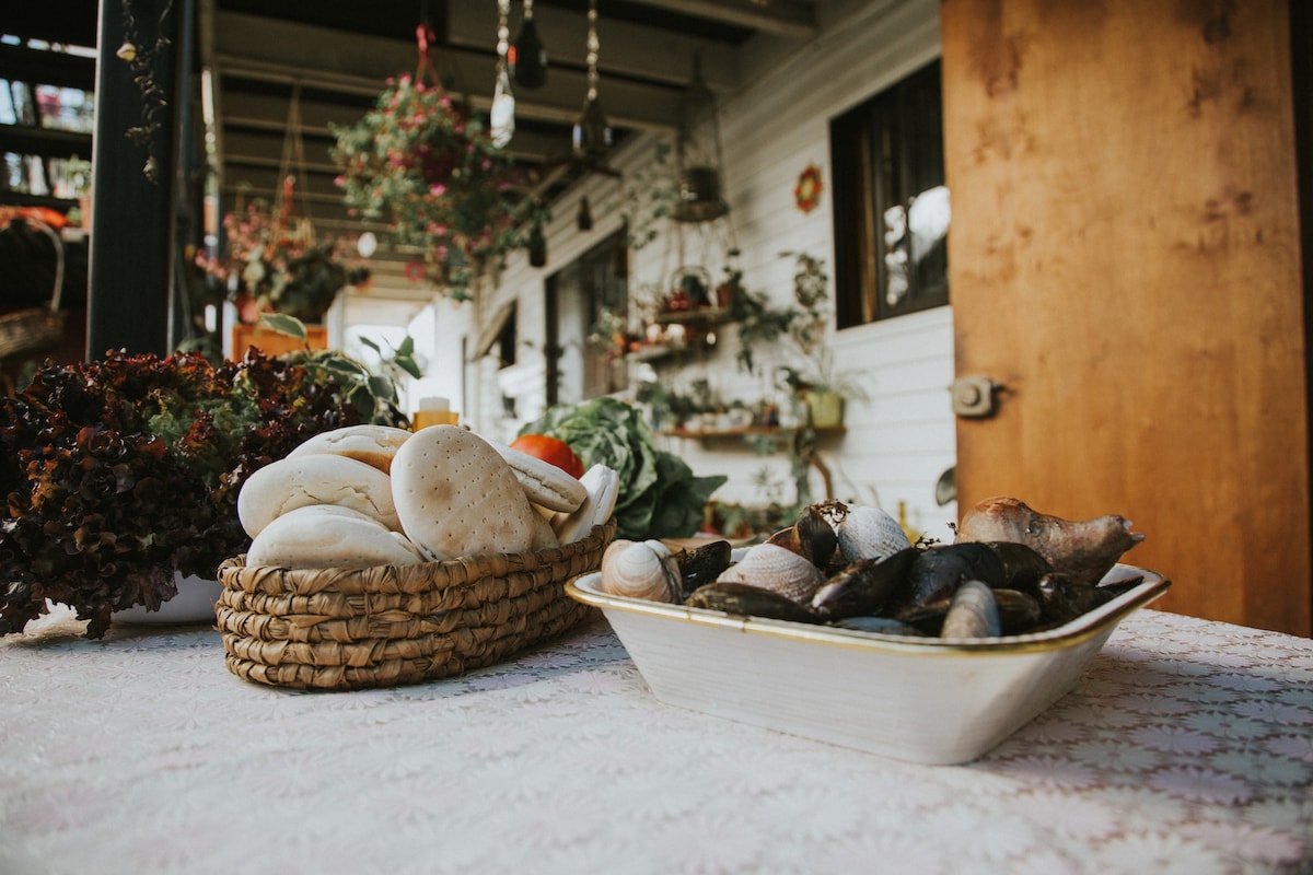 A large basket filled with an assortment of fresh mushrooms and vegetables sits on a lace-covered table. Nearby, a white dish holds an array of shellfish, offering a glimpse of local cuisine. Decorative plants are seen hanging in the background.