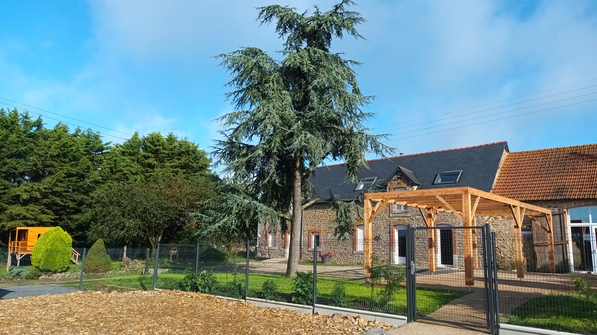 A spacious outdoor area is showcased, featuring a large cedar tree and wooden gazebo structure. Fenced surroundings include landscaped greenery and gravel paths, leading to a charming stone building with multiple windows, under a blue sky with scattered clouds.