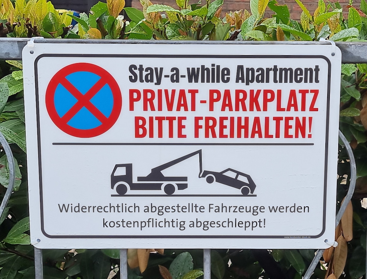 A sign is displayed in front of a private parking area, indicating that the parking space is reserved. The sign features a blue 'X' symbol and includes both German and English text, detailing conditions for parking and towing.