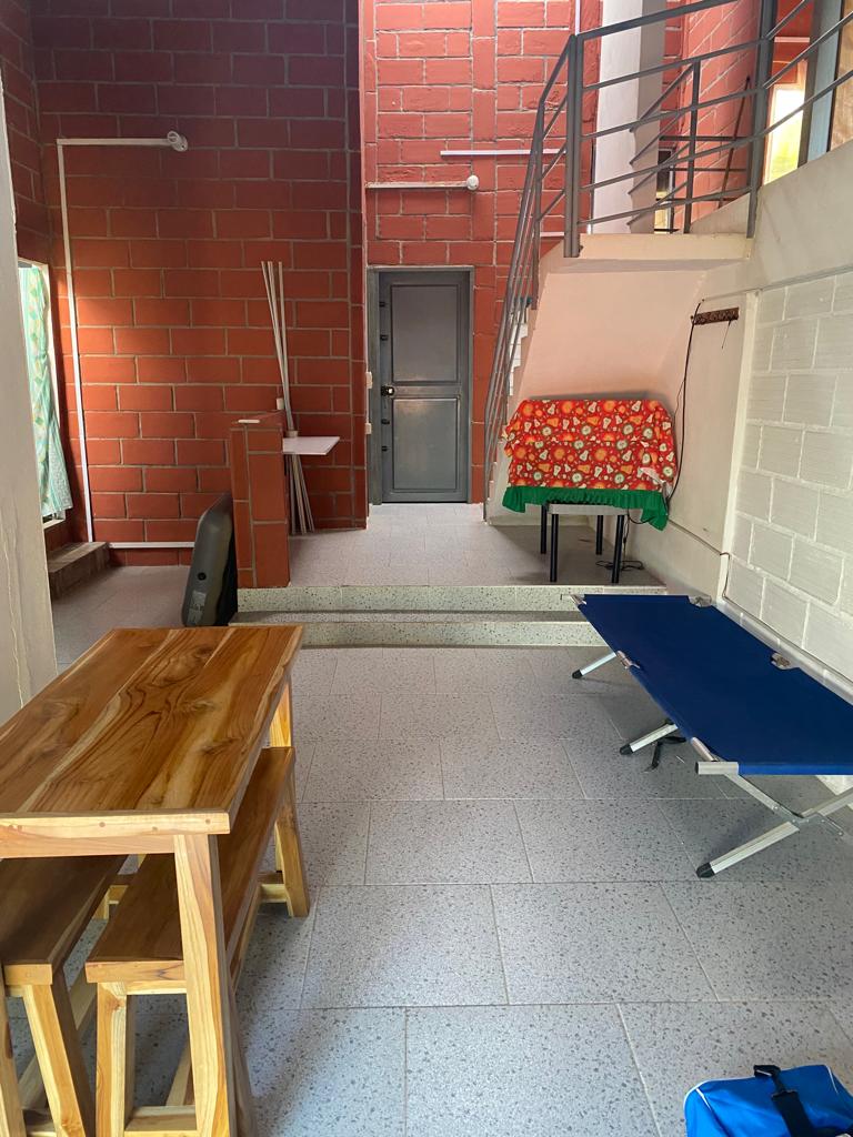 An open space is shown with a wooden table and two stools. A blue folding bed is positioned nearby, and a patterned cloth covers a small table. A door leads to another area, with natural light entering from large windows.