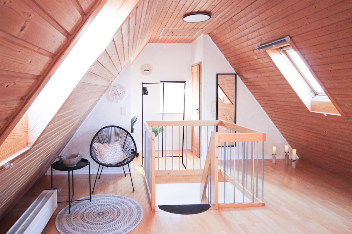 An airy loft area features angled wooden ceilings and natural light streaming through skylights. A round area rug enhances the space, complemented by a decorative chair and a small side table. A wooden railing overlooks the lower level, while candles provide a subtle touch of warmth.