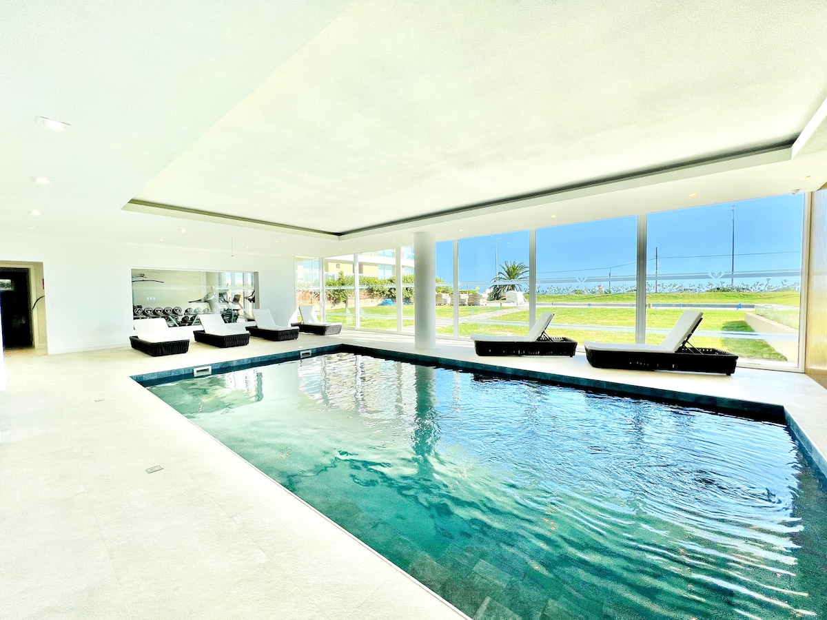An indoor heated pool is surrounded by large glass windows that provide expansive views of the outdoors. Loungers are positioned beside the water, creating a serene space for relaxation. The pool area features light-colored flooring and a clean, modern design.