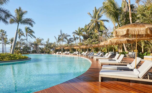 A tranquil pool area is surrounded by lush palm trees, with lounge chairs placed along the wooden deck. Thatched umbrellas provide shade to guests. The gentle curve of the infinity pool creates a seamless connection with the clear blue sky.
