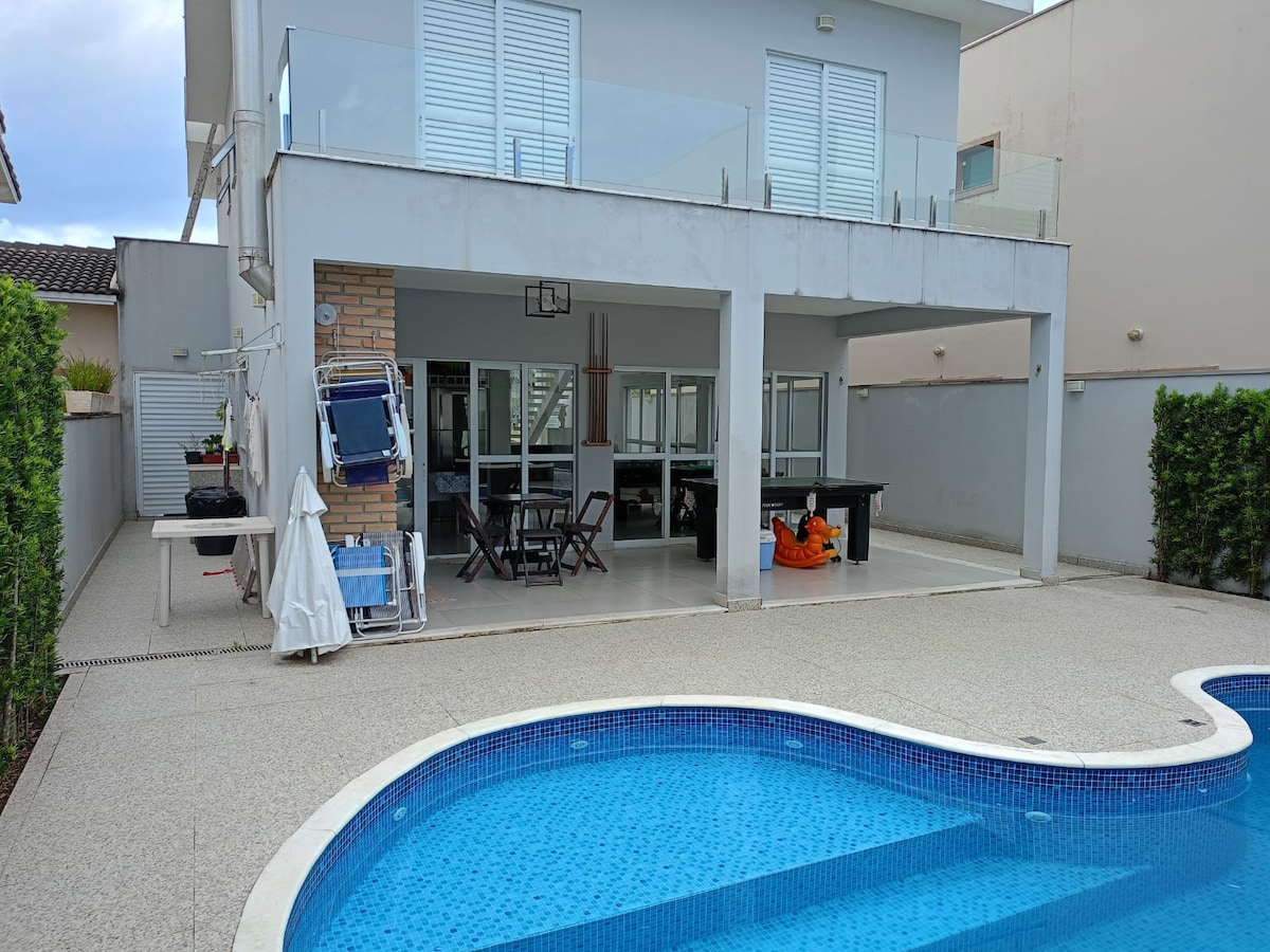 A spacious patio area is visible, featuring a curved pool with a mosaic tile design. Multiple seating areas are arranged around the pool, with a dining table and pool table situated under a covered area. The building's large windows provide a view into the interior.