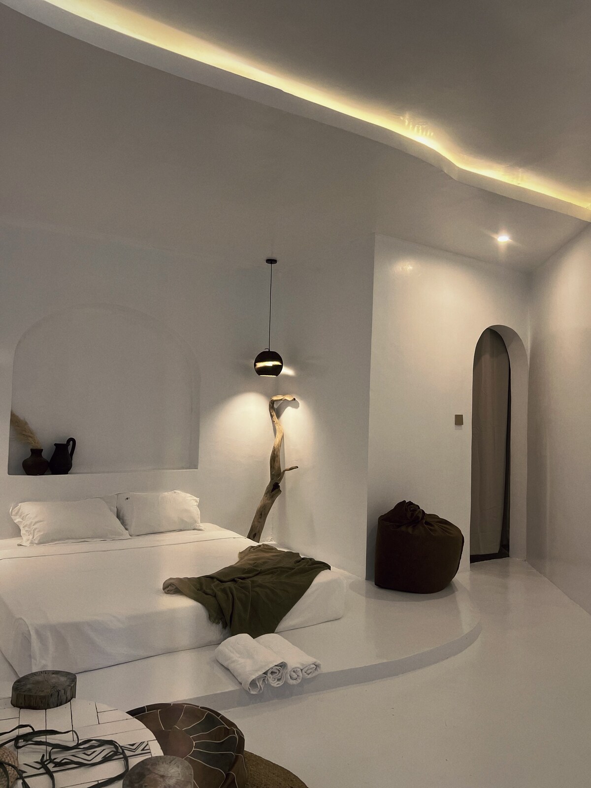 A clean, minimalist bedroom features a spacious bed dressed in soft linens. Natural elements are included with a tree branch accent and a light fixture. A cozy seating area is highlighted by a large, round pouf and decorative textiles, complementing the serene atmosphere.