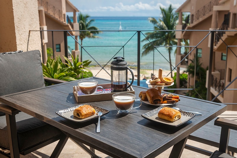 A dining table is set for breakfast on a balcony overlooking the beach, featuring coffee, pastries, and a french press. Lush greenery and a glimpse of the infinity pool are visible in the background against the serene ocean view.