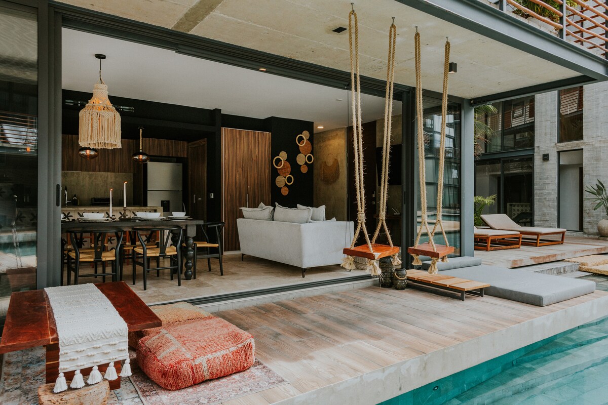A modern living space is highlighted by large glass doors opening to a patio, featuring a swimming pool. Soft seating areas and decorative rugs are arranged around the outdoor space, while stylish swings hang nearby, creating a relaxed environment for gatherings.