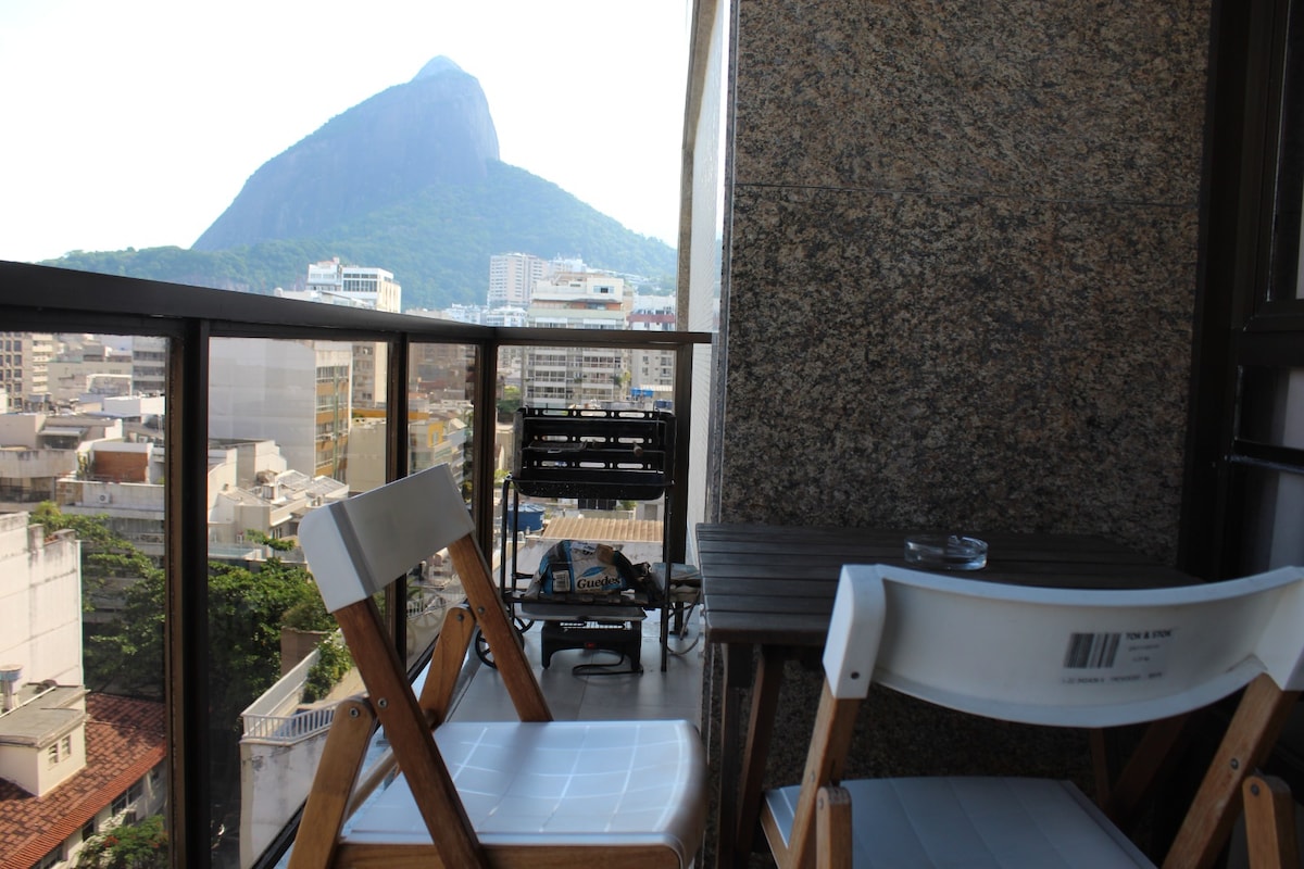 A balcony scene features a small wooden table accompanied by two white chairs. In the background, the iconic Morro Dois Irmãos is visible, surrounded by a vibrant urban landscape with rooftops and buildings.