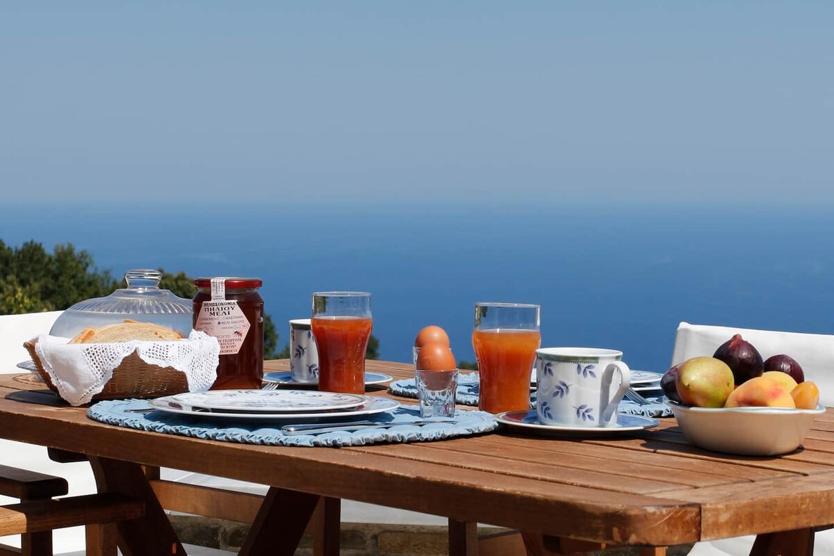 A wooden dining table is set outdoors, featuring an arrangement of breakfast items. Glasses filled with juice, plates with fruit, and a jar of jam are visible. A view of the expansive sea is seen in the background, contributing to a serene setting.