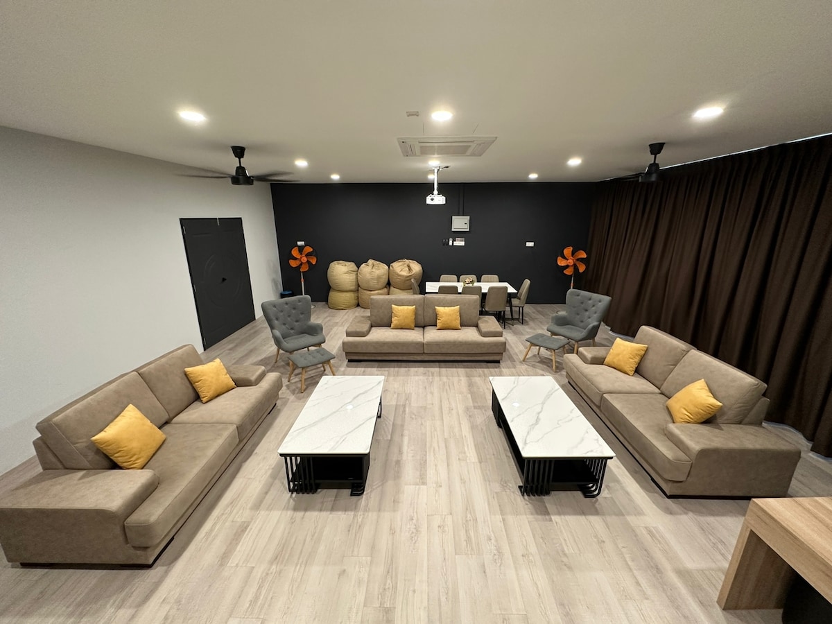 A spacious living area features comfortable seating with neutral-toned sofas adorned with yellow cushions. Two marble-topped tables are positioned in the center, and the background includes a dark wall with subtle decor elements. Ceiling fans and warm lighting enhance the inviting ambiance.