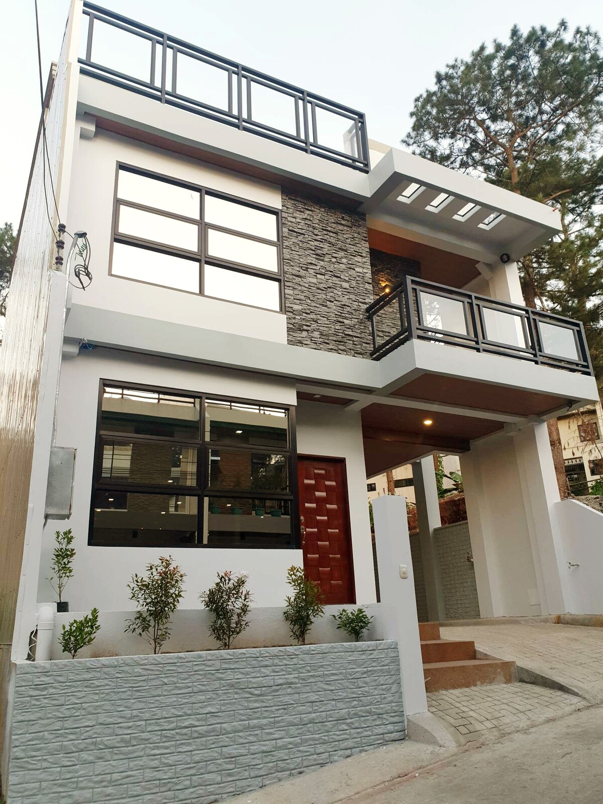 A modern vacation house is displayed, featuring a combination of stone and smooth exterior finishes. Large windows provide ample natural light, and a welcoming front entrance is complemented by simple landscaping with low shrubs and a pathway leading to the steps.