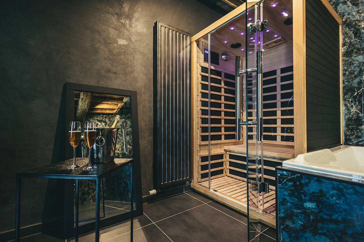 A modern sauna with illuminated interior and wooden slats is featured. A sleek mirror reflects the space, while a small table holds two flutes and a champagne bottle. The room's dark tones add to a serene atmosphere, complemented by accent lighting.