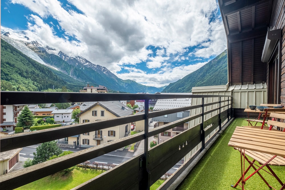 A balcony is shown with outdoor seating options, featuring two wooden chairs and a table. The view includes a scenic landscape of mountains and a valley under a partly cloudy sky, enhancing the connection to nature.