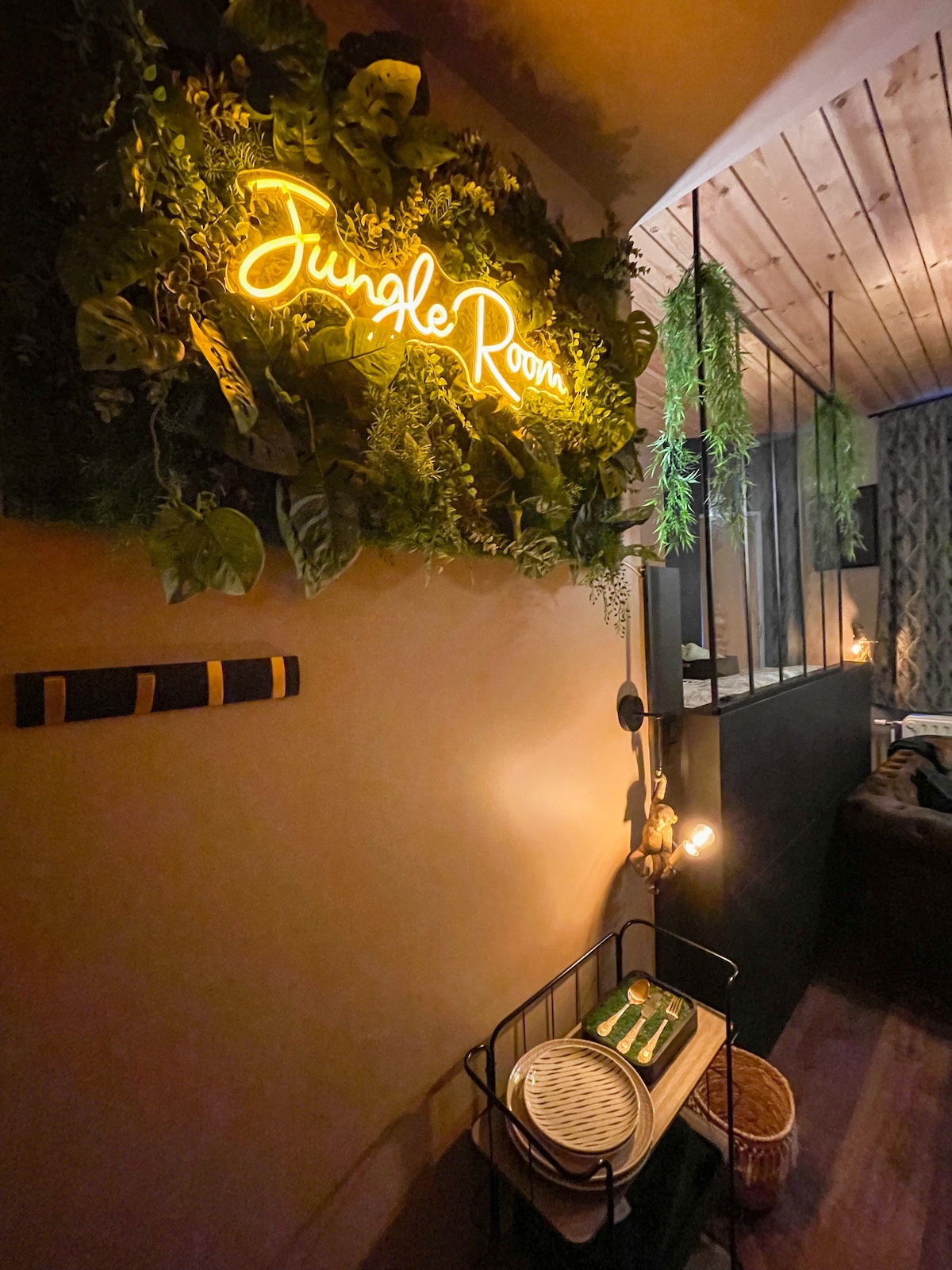 An inviting entrance area is featured, characterized by a vibrant 'Jungle Room' neon sign. Lush greenery adorns the wall, while warm lighting creates a cozy ambiance. A decorative basket is placed on a stand, contributing to the room's overall charm.
