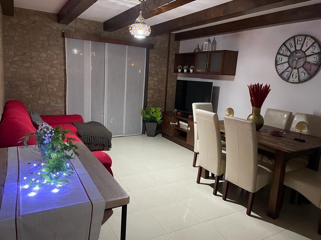 A bright living space features a red sofa and a wooden dining table surrounded by light-colored chairs. Natural light enters through sheer curtains, illuminating the room. Decorative elements include a clock on the wall and a plant near the television, enhancing the welcoming environment.