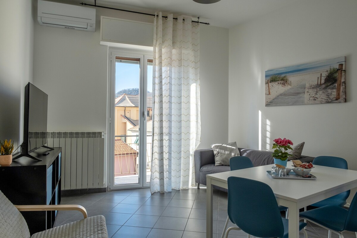 A bright living area is presented with a comfortable sofa and a dining table surrounded by chairs. A large window offers natural light, complemented by sheer curtains. A television is positioned on a cabinet, and a coastal-themed artwork decorates the wall.