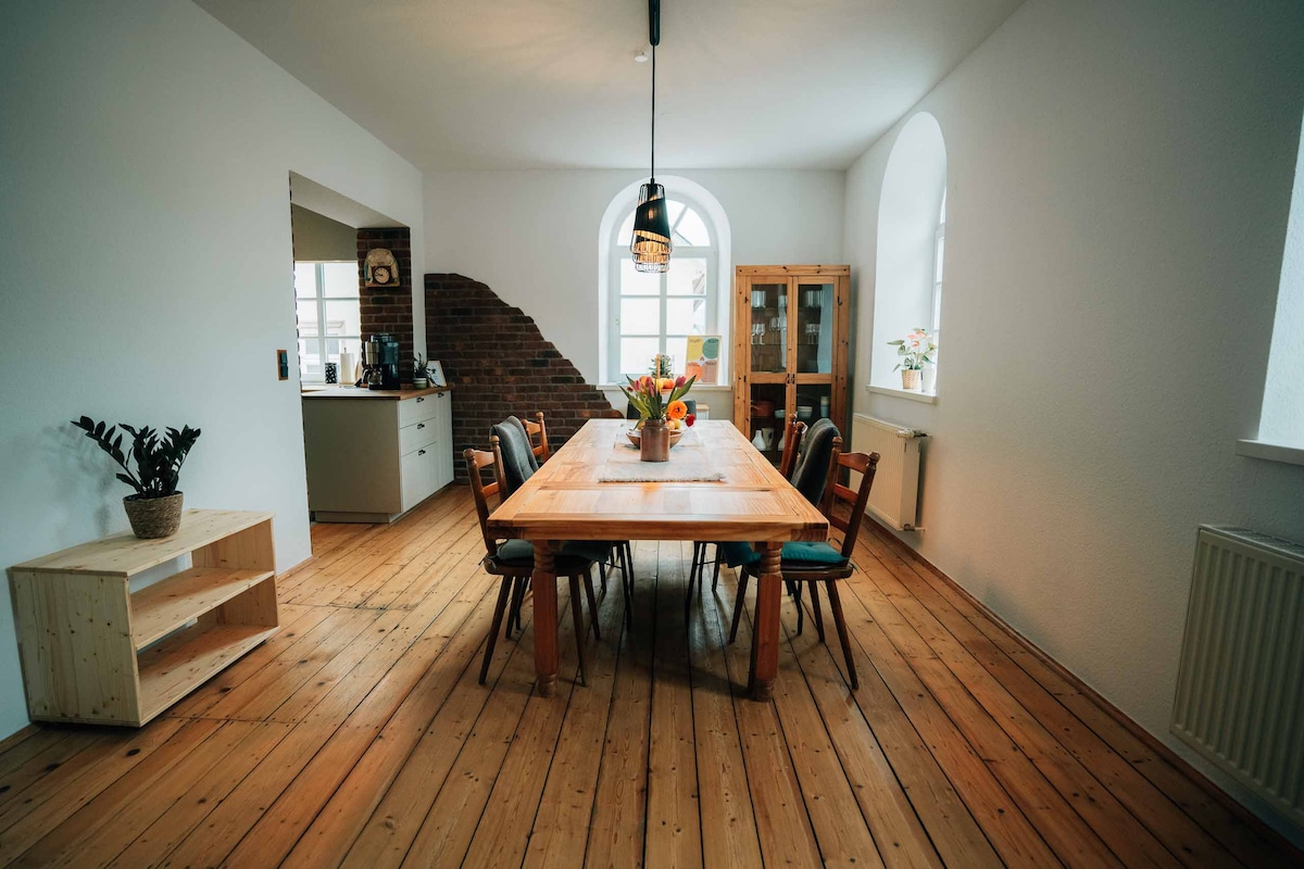 A bright dining area features a long wooden table surrounded by chairs, accommodating casual meals. Large arched windows provide natural light, and a decorative vase filled with flowers sits at the table's center. A small side table adds to the room's functionality.