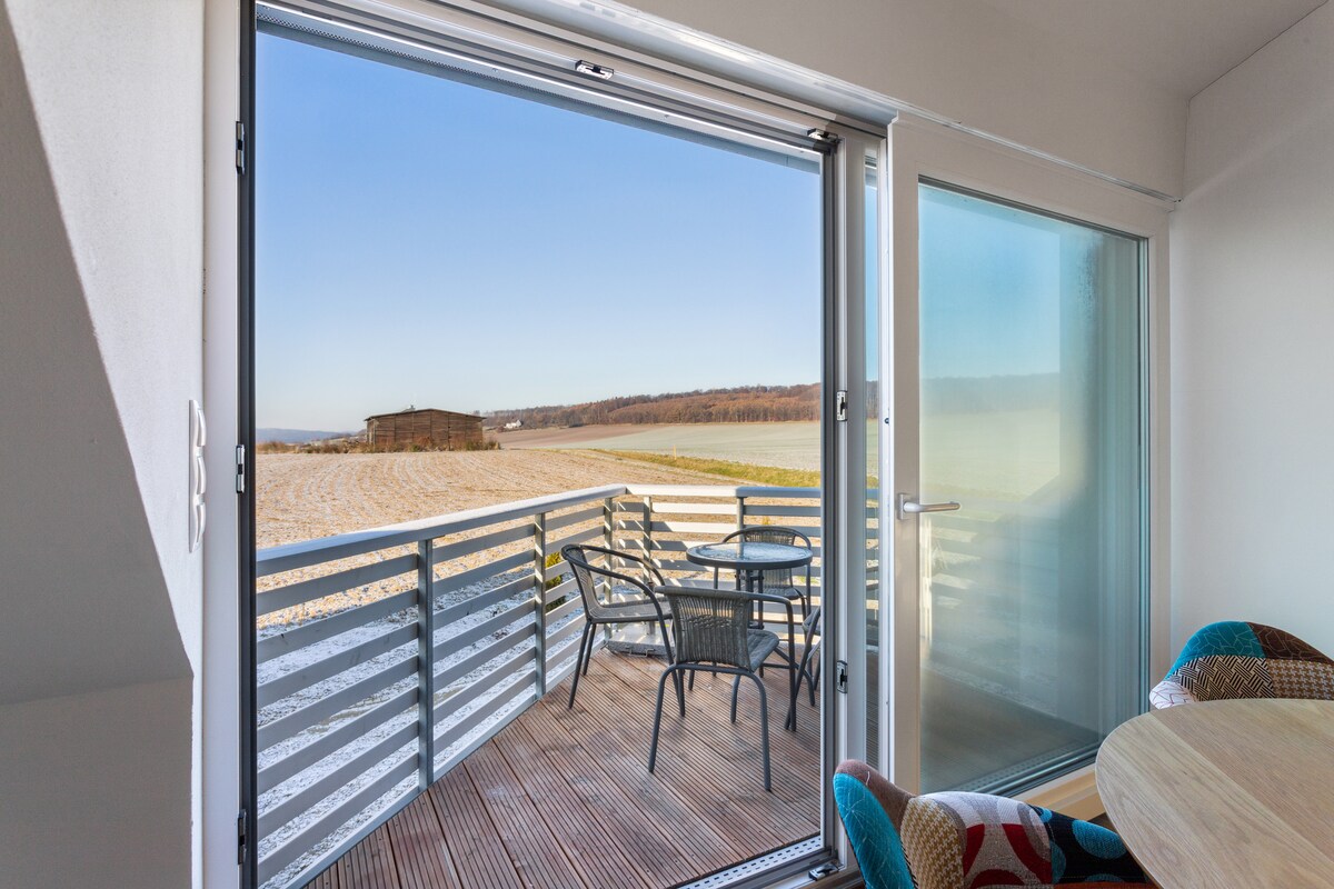 An outdoor balcony features a table and chairs, offering views of a serene landscape. The sliding glass doors provide easy access, enhancing the connection with the natural surroundings.