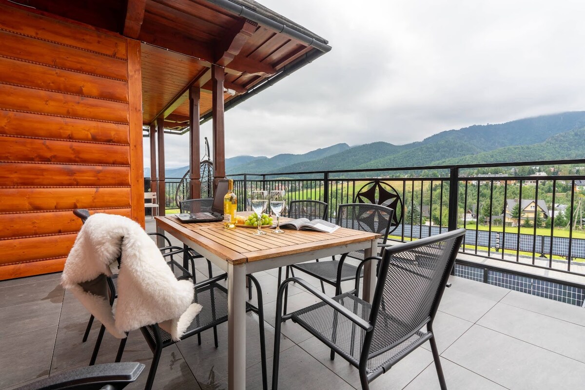 An outdoor terrace features a wooden table surrounded by four black chairs, with a scenic view of rolling mountains in the background. A bottle of wine and glasses are placed on the table, inviting relaxation. The space is partially covered by a wooden roof, providing shelter.
