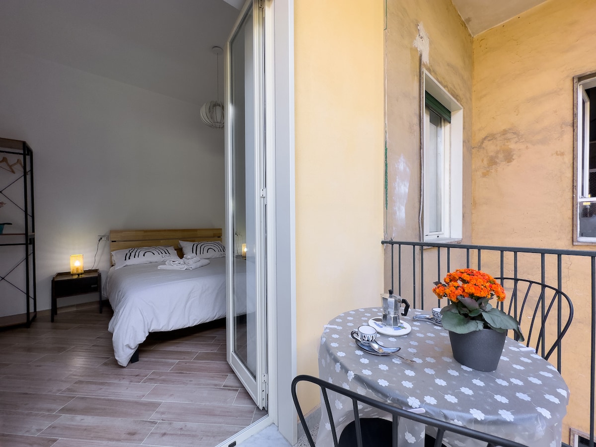 A small outdoor space features a round table set for two, with a potted orange flower centerpiece. In the background, a spacious bed is visible inside the room, complemented by subtle lighting on nightstands. Large doors open to connect the indoor and outdoor areas.