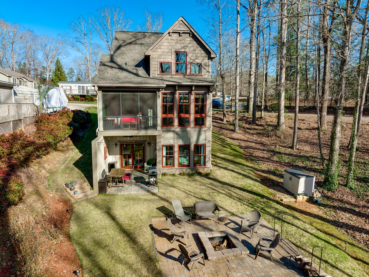 The exterior of the property showcases a spacious backyard featuring a stone patio surrounded by seating areas and a fire pit. The house, with large windows, is set against a backdrop of trees, providing a serene outdoor environment.