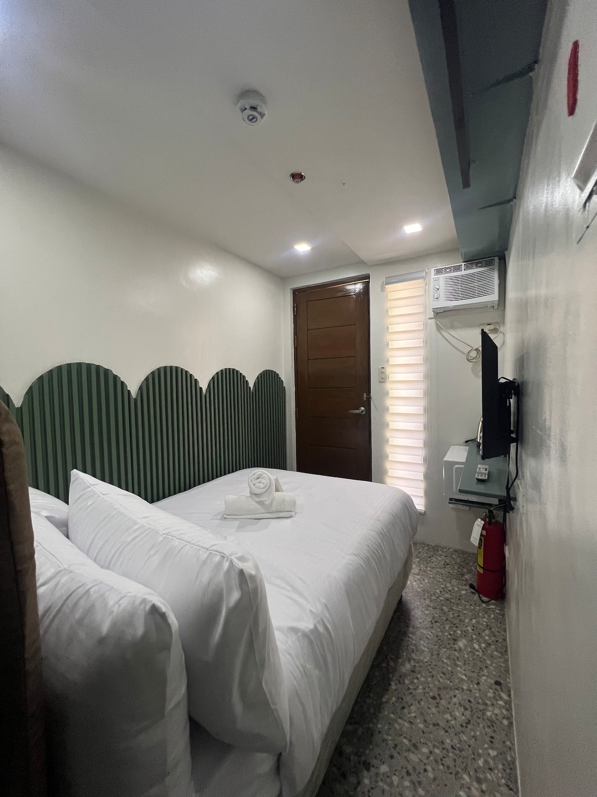 A compact sleeping area features a spacious bed with crisp white linens and a decorative green accent wall. A flat-screen television is mounted near the foot of the bed. Natural light filters in from a window covered with blinds, and a wooden door leads to the corridor.