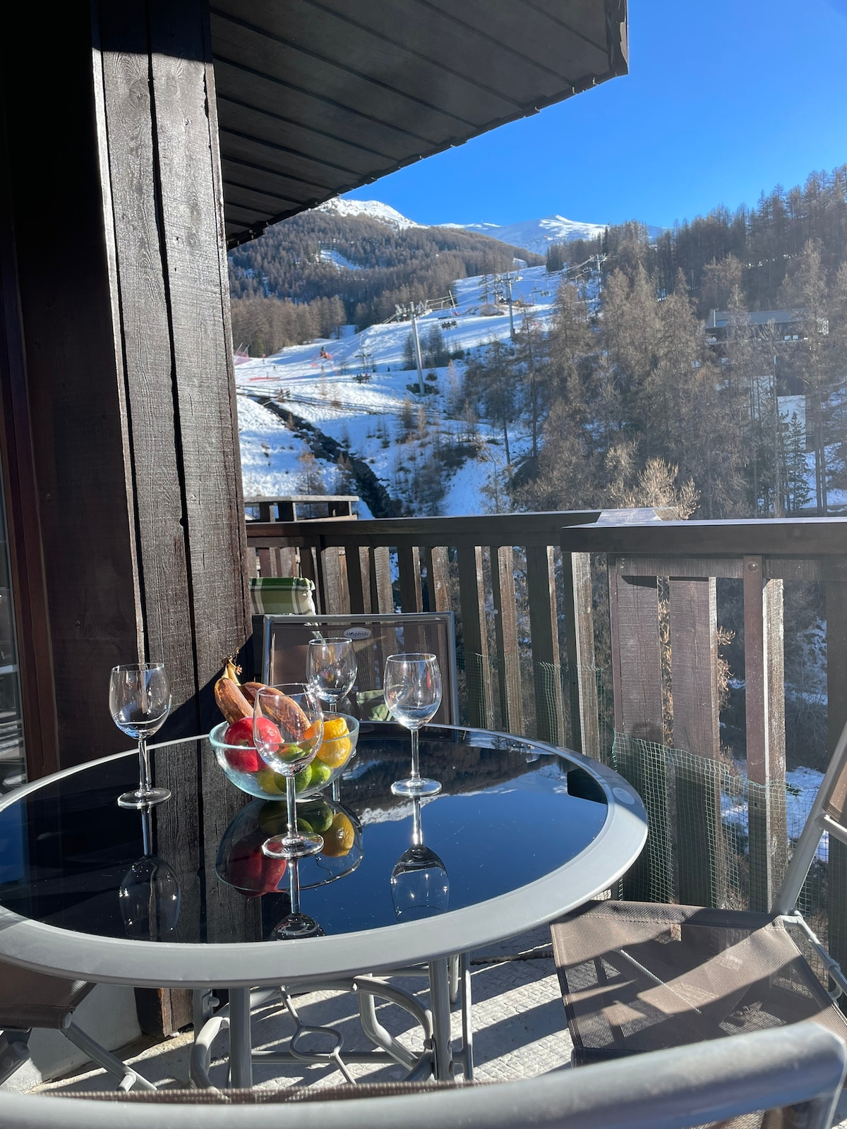 A balcony table is set with glasses and a bowl of fresh fruit, providing a view of snow-covered slopes and pine trees in the distance. The bright blue sky enhances the scenic mountain backdrop, creating a serene outdoor space.
