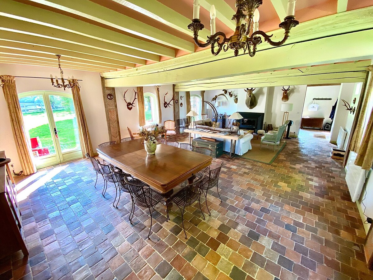A spacious living area features a long wooden dining table surrounded by wicker chairs, complemented by rustic wooden beams overhead. A seating arrangement is positioned near a fireplace, and large doors open to nature, enhancing the connection to the outdoor space.