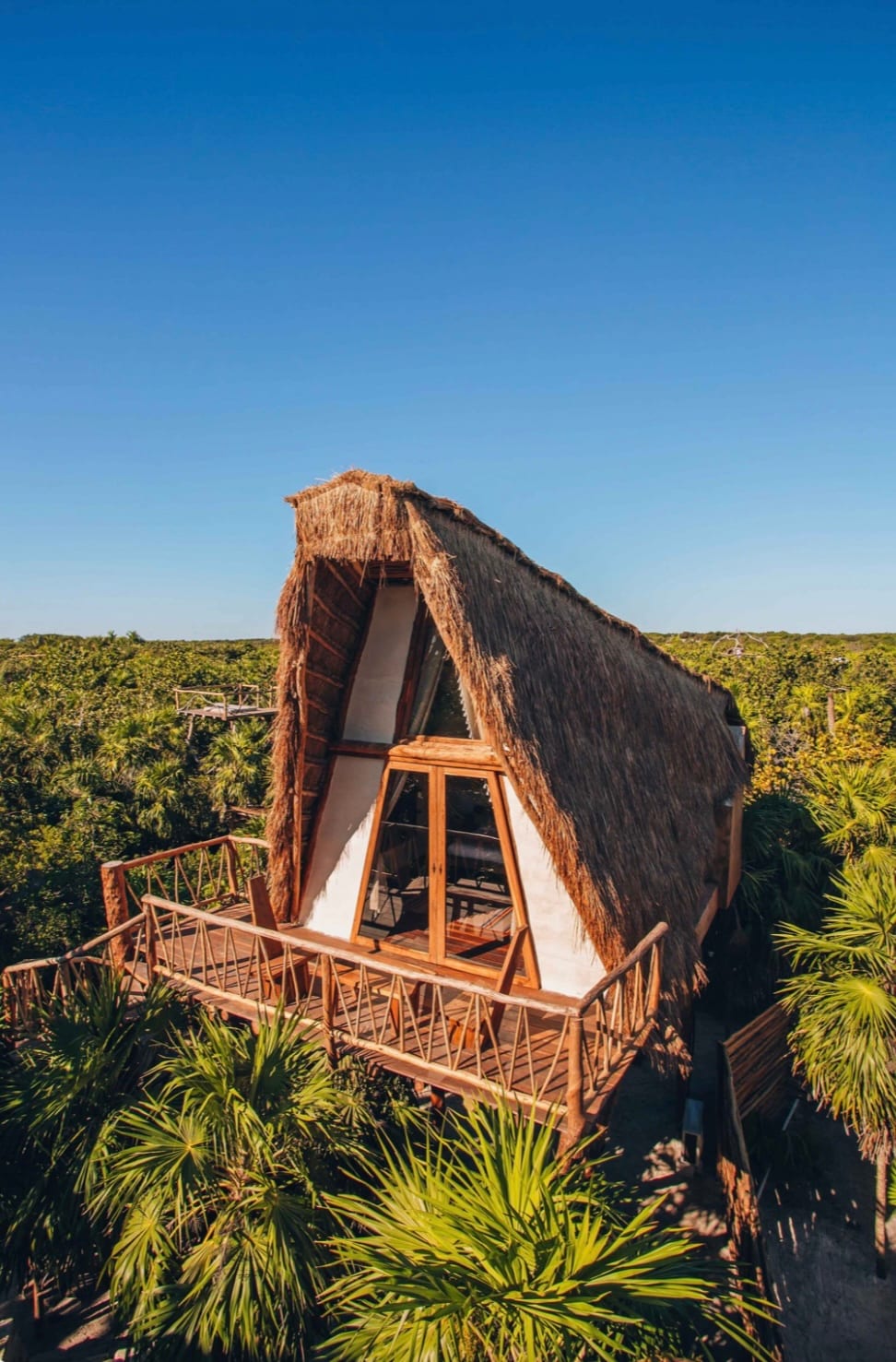 A charming treehouse suite is positioned atop a raised platform, featuring a thatched roof and wooden balcony. Surrounded by lush greenery, large windows integrate natural light, while the expansive deck provides a peaceful spot for relaxation amidst the treetops.