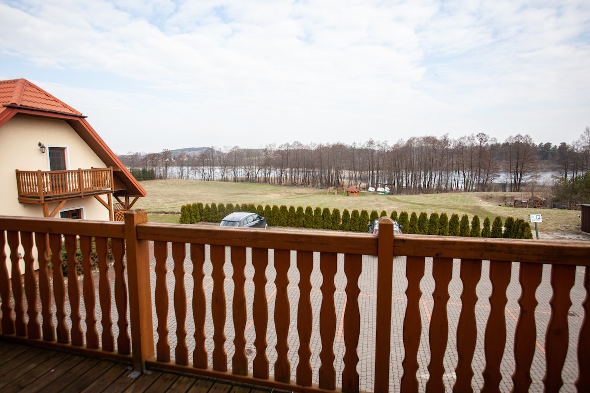 A wooden balcony railing offers an unobstructed view of a serene landscape, featuring a lake and grassy areas. In the distance, trees line the horizon under a partly cloudy sky. A vehicle is parked nearby, adding to the peaceful scene.