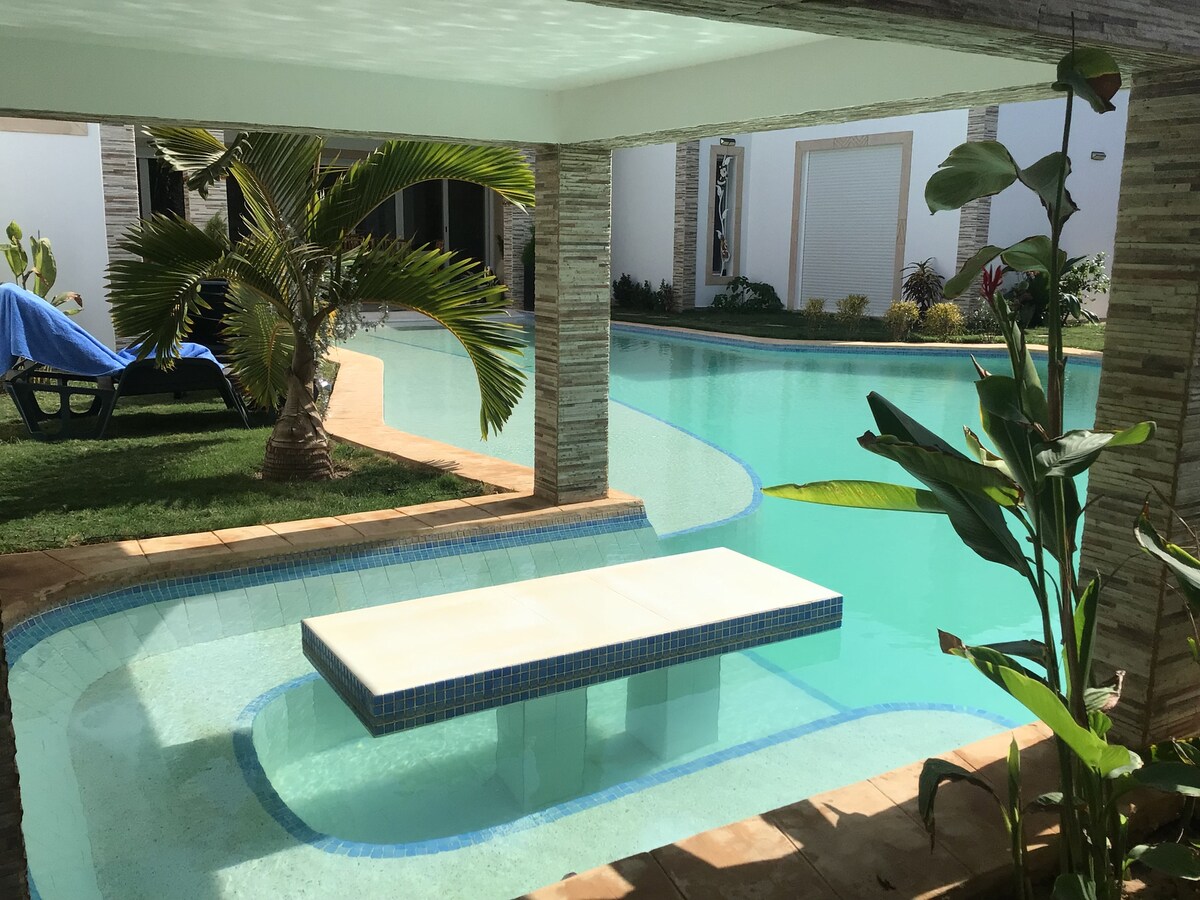A serene outdoor space features a shimmering blue pool that curves around a central platform. Lush green plants are positioned nearby, while a sunbed can be seen to the left, inviting relaxation in the tranquil setting.