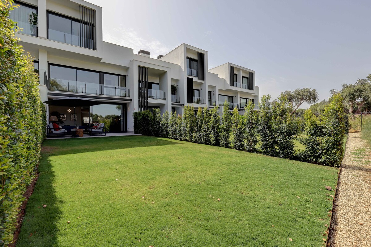 The modern townhouse is complemented by a spacious lawn, lush with green grass. Large windows reflect natural light and create a sense of openness. The surrounding hedges provide privacy, framing the home and enhancing the outdoor space suitable for relaxation or socializing.