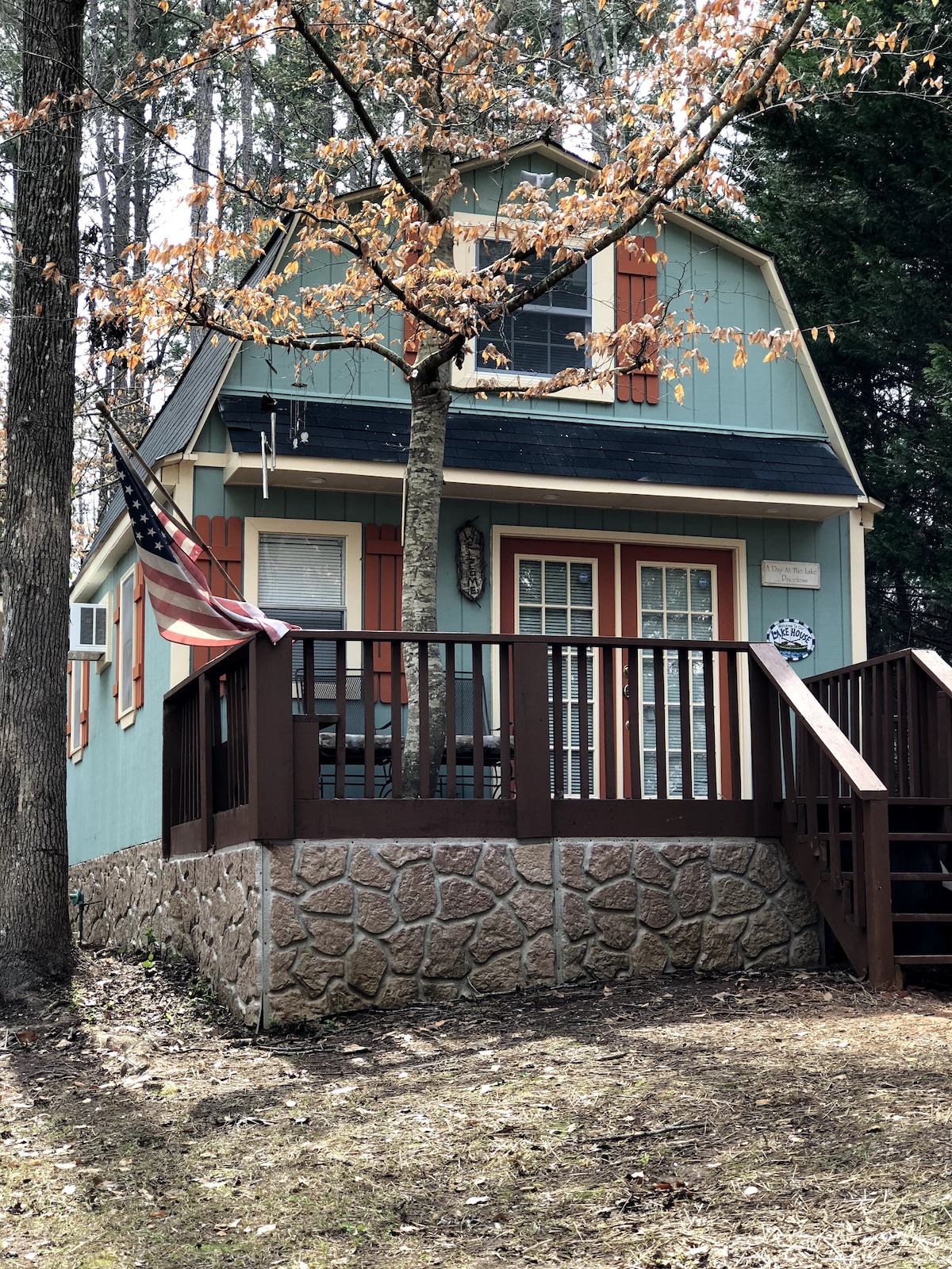 A charming lake house features a light blue exterior complemented by orange shutters. A small set of stairs leads to a welcoming porch, where an American flag hangs. Surrounding trees add a natural touch to the peaceful setting.