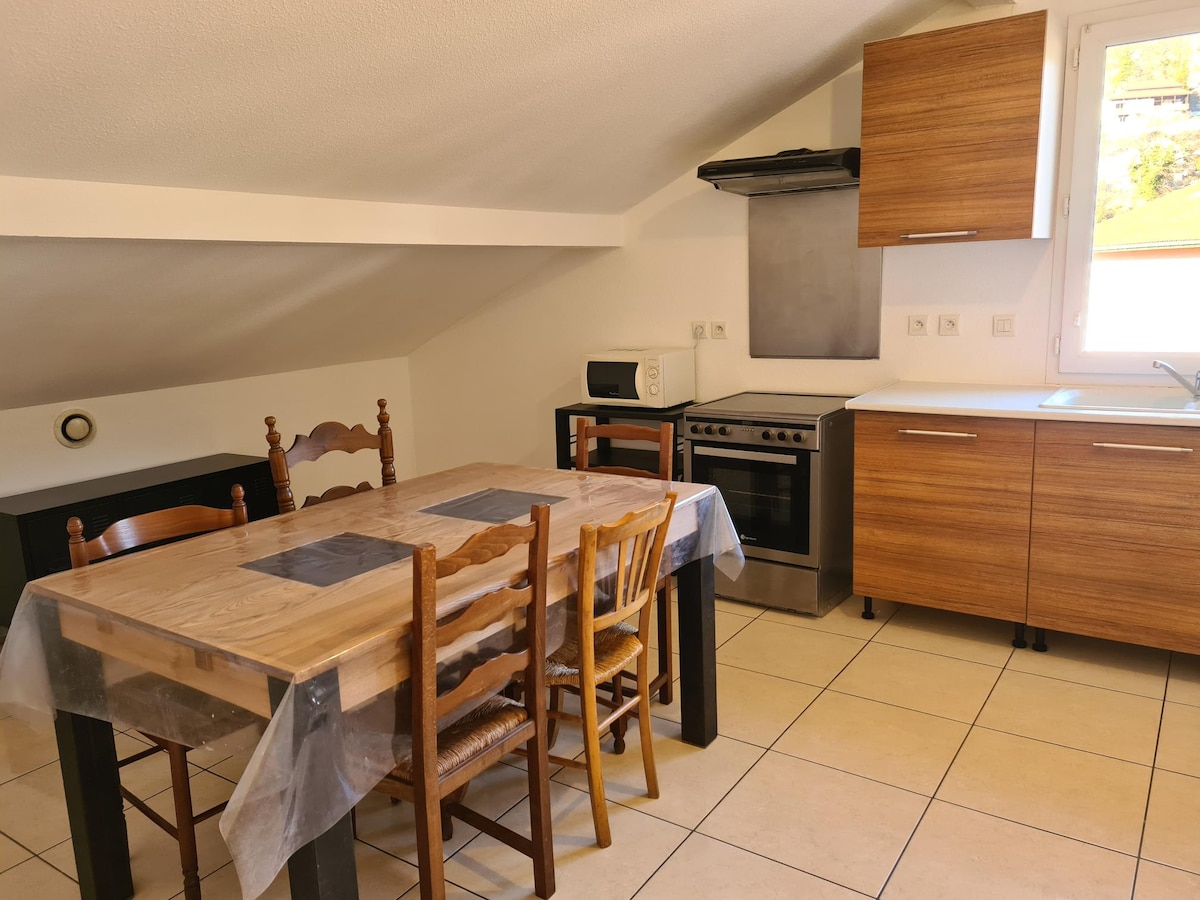 A well-organized kitchen features wooden cabinets and a stove with an overhead extractor. A dining table with six wooden chairs is positioned in the center. A microwave and other kitchen appliances are visible, with light streaming in from a nearby window.