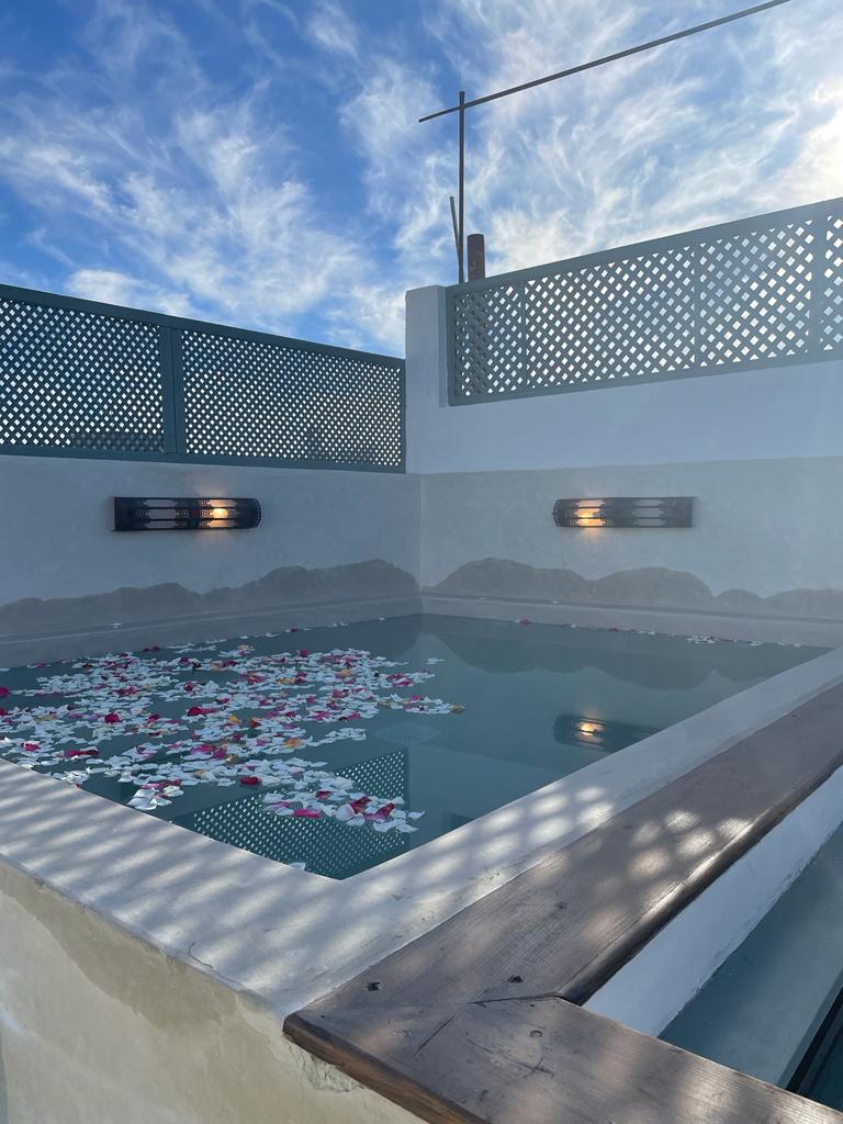 A serene private pool is surrounded by textured walls and wooden accents. The water's surface is adorned with floating petals in various colors. Softly lit wall sconces provide gentle illumination, while a lattice screen allows light to filter through, creating a tranquil ambiance.
