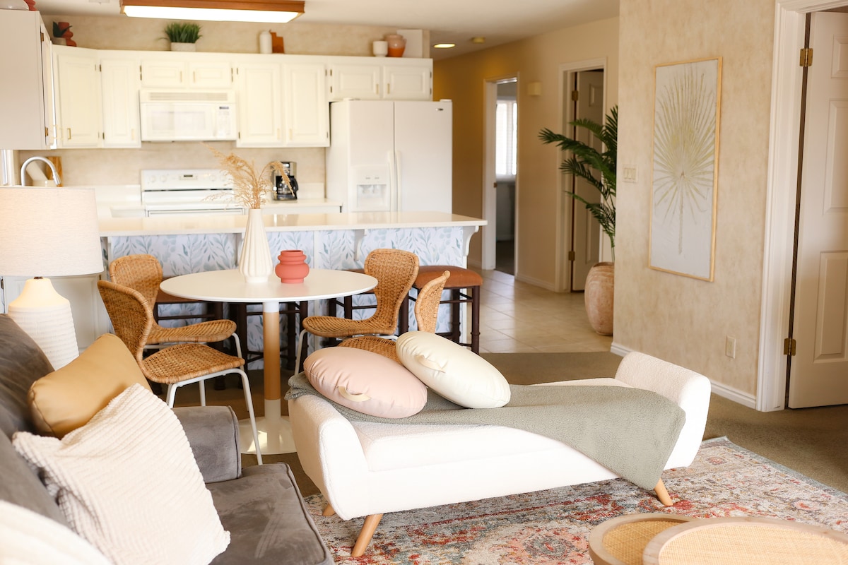 An open living area features a comfortable couch adorned with pillows and a textured rug. A round dining table is surrounded by wicker chairs, while the kitchen is visible in the background, showcasing white cabinetry and appliances. Natural light floods the space through large windows.