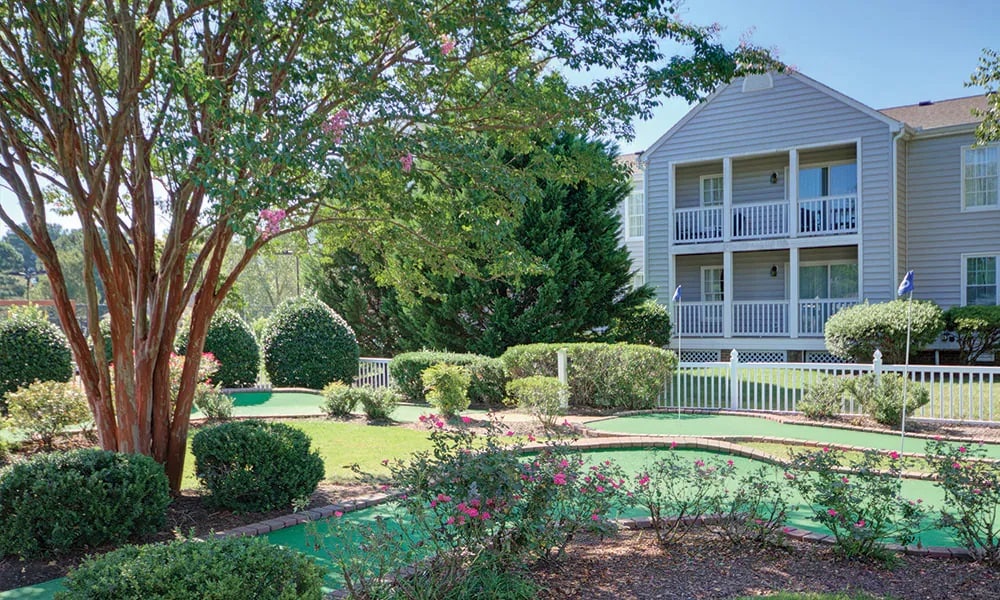 A manicured outdoor area features a miniature golf course surrounded by lush green lawns and flowering bushes. The two-story building in the background displays balconies, while tall trees provide shade over the vibrant landscape.