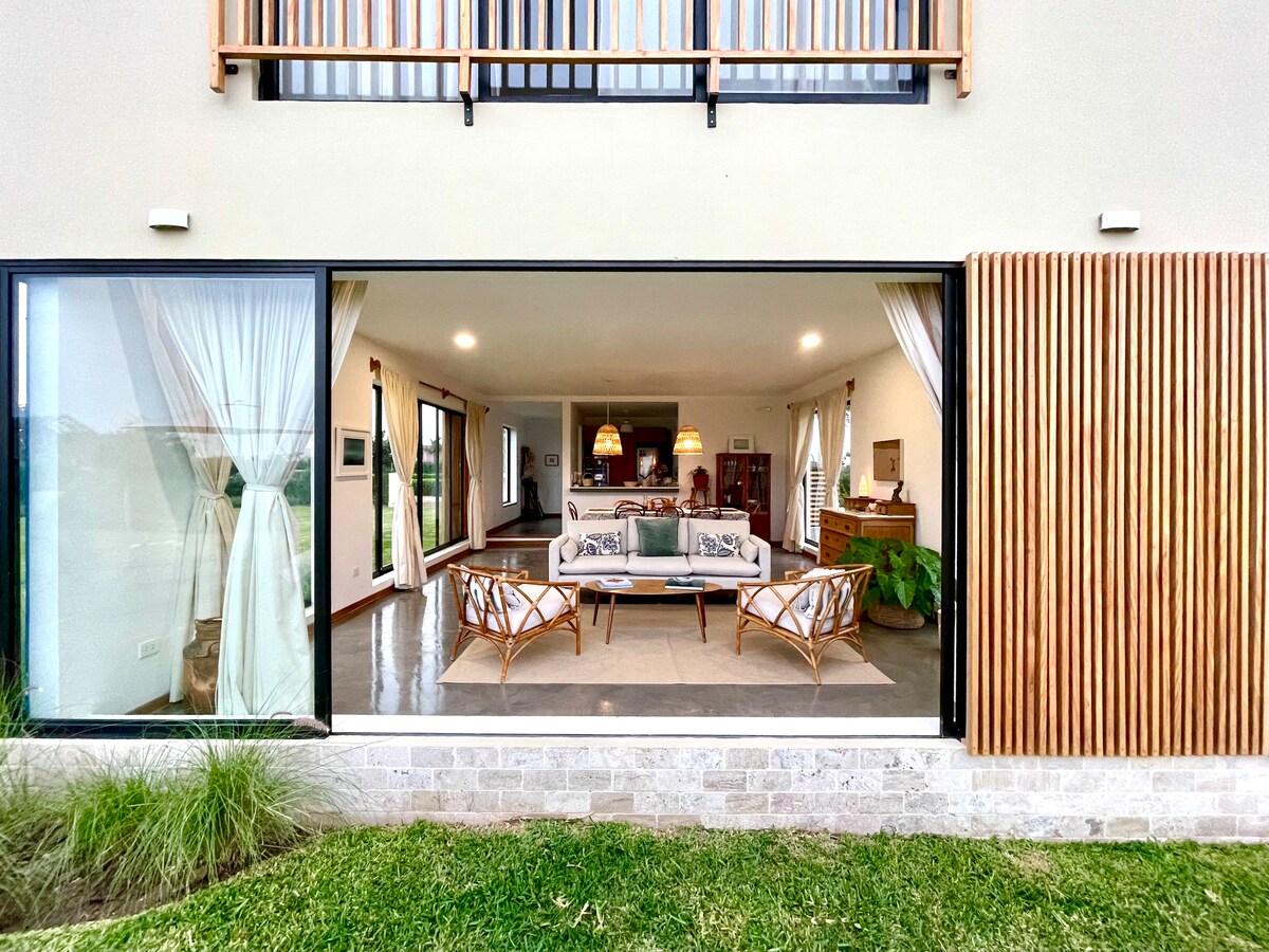 A spacious living room is depicted, featuring large glass doors that open to the exterior. Comfortable seating is arranged around a central coffee table, with natural light illuminating the room. Elegant decor elements are visible, creating a welcoming space that connects indoor and outdoor environments.