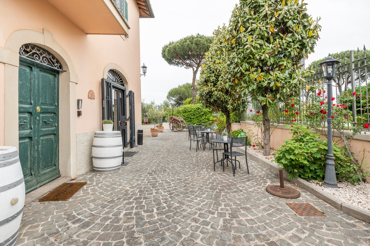 A cobblestone courtyard is framed by a charming building, featuring vintage green doors and an elegant wrought-iron gate. Outdoor seating is provided by black metal tables and chairs, surrounded by greenery and flowering shrubs, creating a serene setting in the countryside.