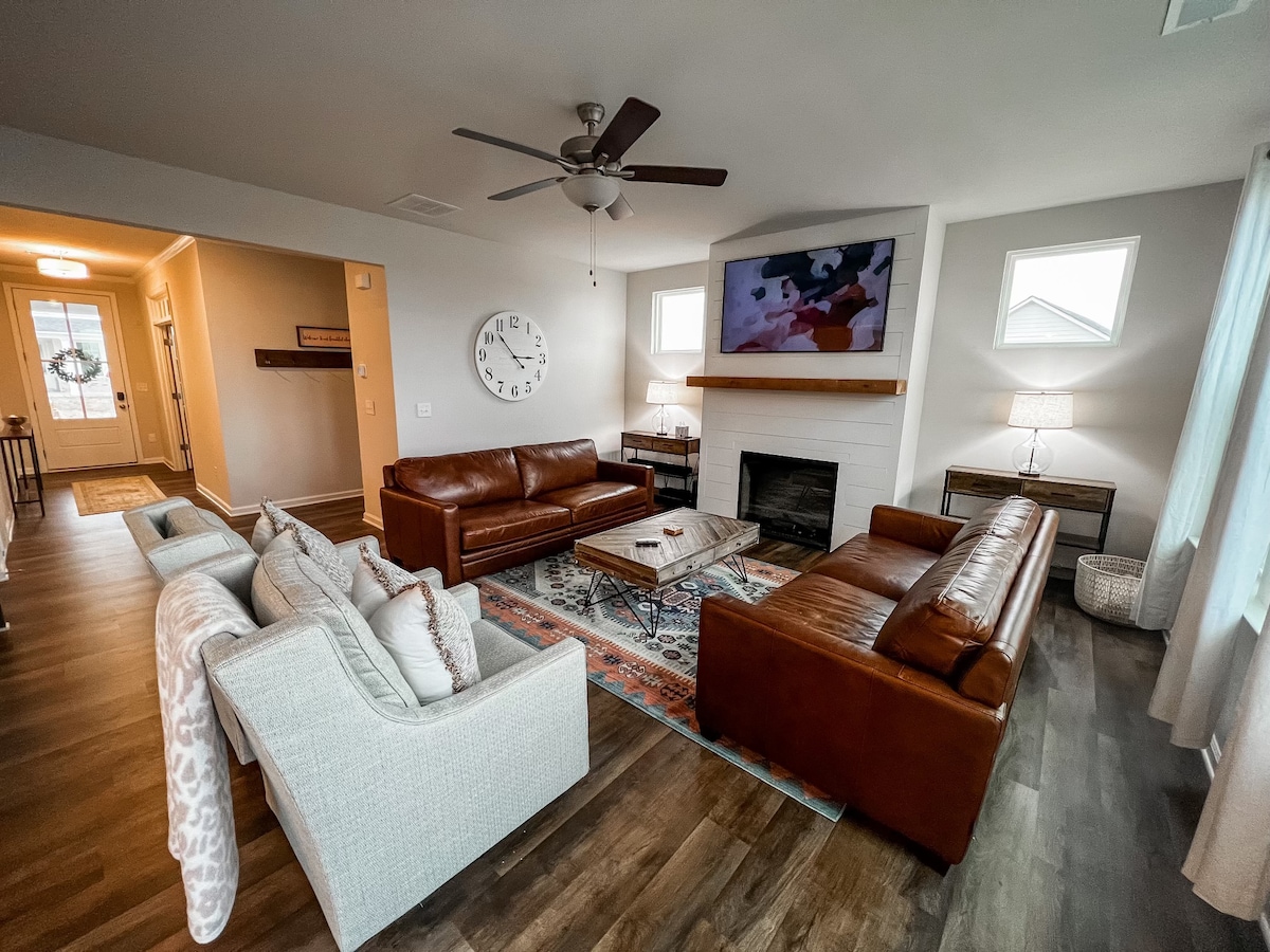 The living room features two brown leather couches and a light-colored couch, arranged around a coffee table. A flat-screen television is mounted above a white fireplace. Light streams in through large windows, and a wall clock complements the modern decor.