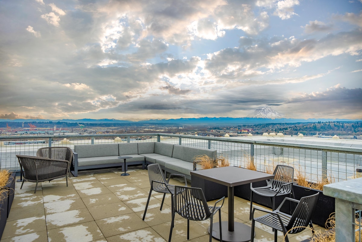 A spacious rooftop terrace offers seating with gray sectional sofas and black chairs, surrounded by panoramic views of Mount Rainier and the city. The paved floor features subtle stains from rain, complementing the natural surroundings of the landscape and sky.