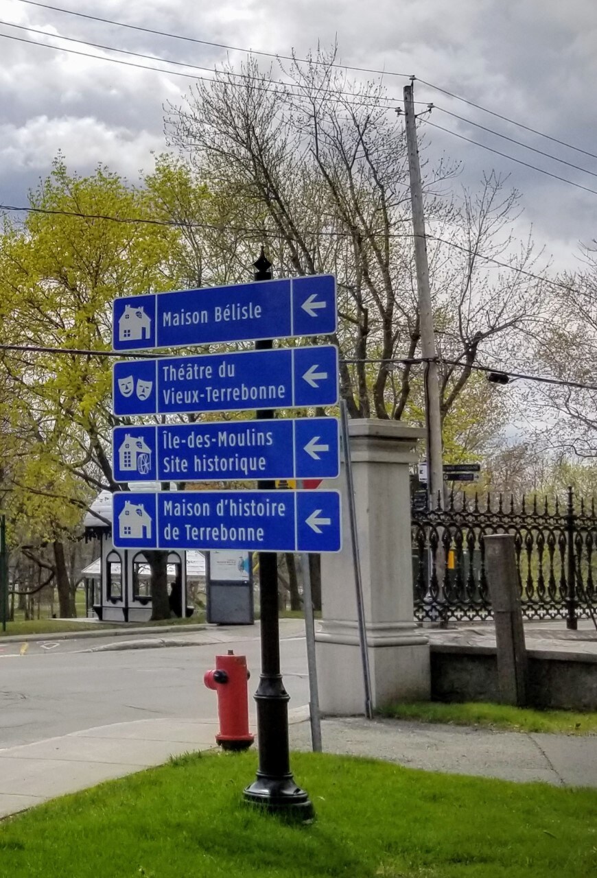 A directional signpost displays various blue signs indicating local attractions. The signs point towards Maison Bélisle, Théâtre du Vieux-Terrebonne, Île-des-Moulins Site Historique, and Maison d'histoire de Terrebonne, amidst a backdrop of trees and a residential area.