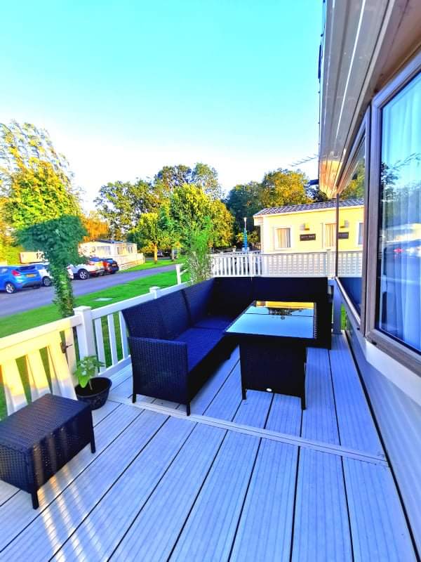 A patio area is present, featuring a black outdoor seating arrangement around a glass-top table. Surrounding greenery enhances the space, while well-maintained decks provide a comfortable setting. Nearby, other caravans and landscaped greenery are visible, contributing to a relaxed outdoor atmosphere.